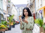 Young smiling Indian woman walking in the city, woman holding a bank credit card and phone, tourist making online booking of accommodation and booking tourist services while walking in the city.